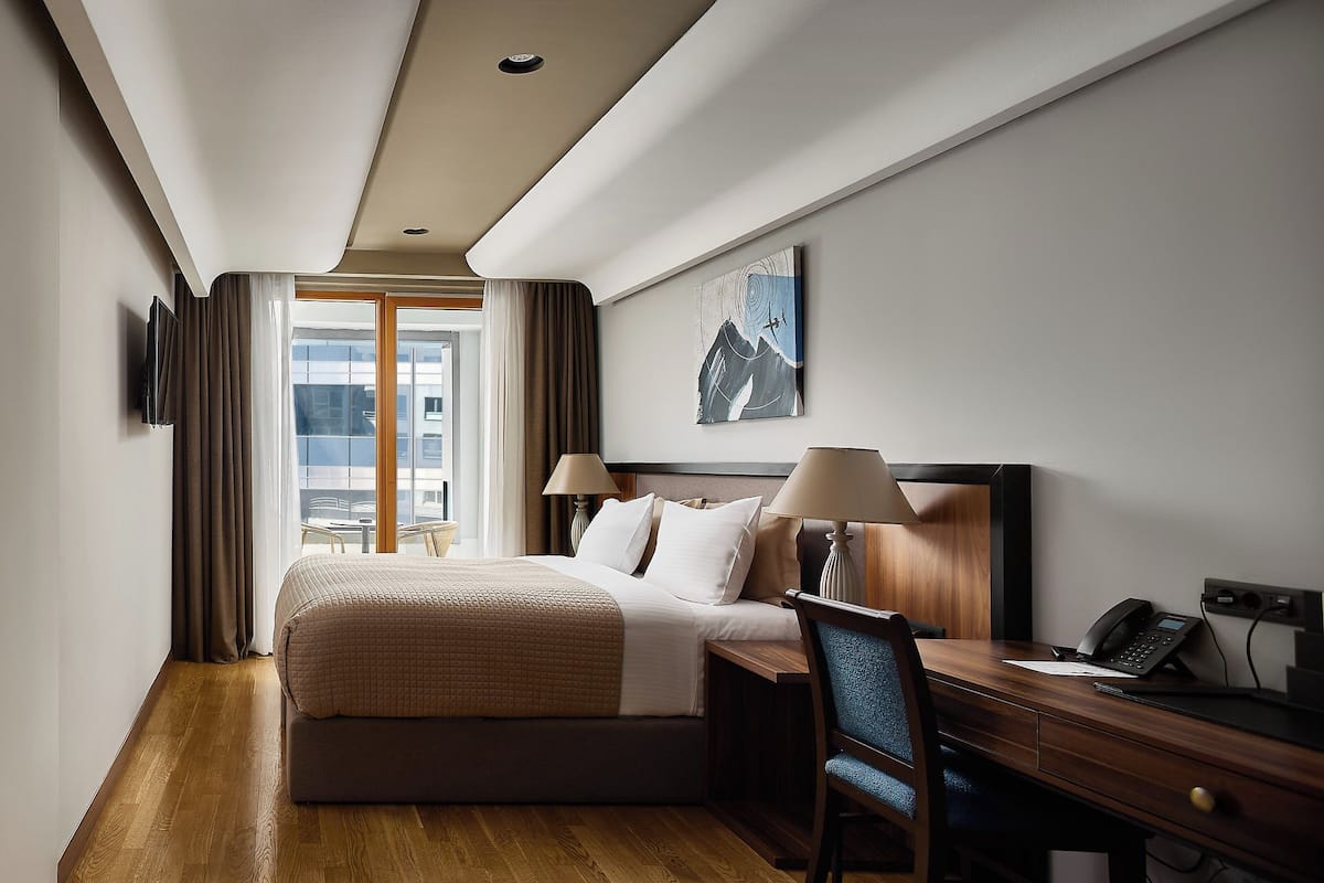Ein Hotelzimmer mit einem großen Bett, einem Schreibtisch mit Telefon und Blick auf die Stadt durch das Fenster.