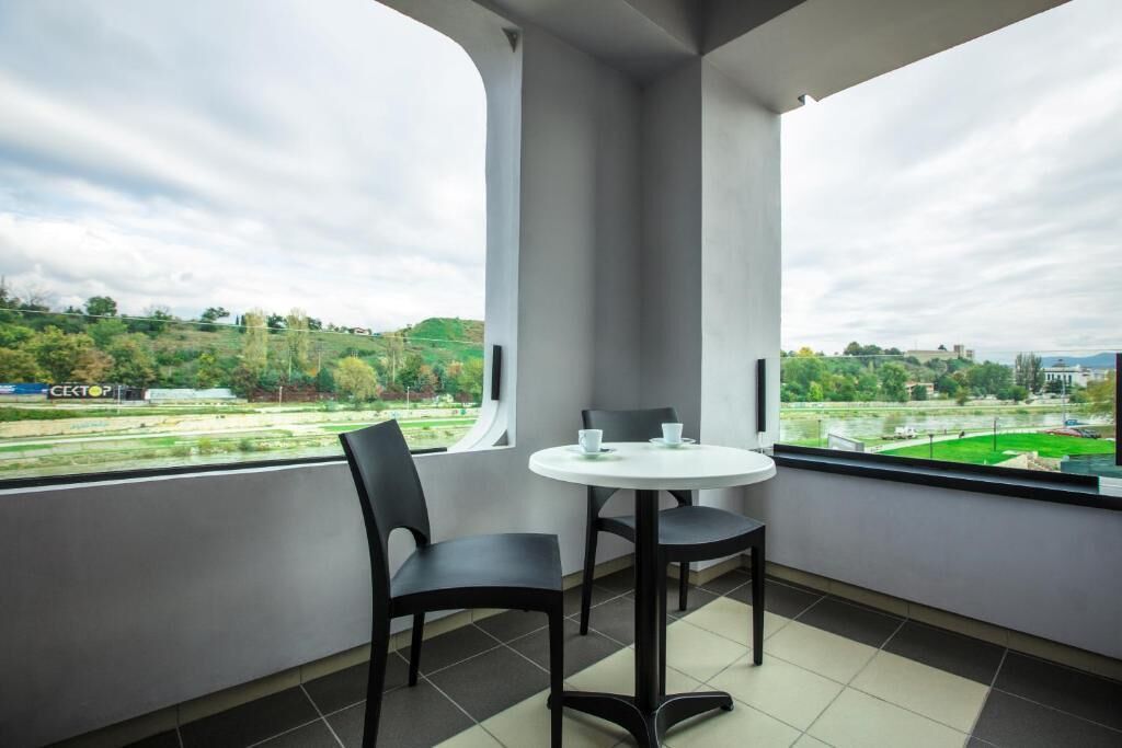 Un balcon avec une table ronde et deux chaises, une grande fenêtre donnant sur un paysage verdoyant et des bâtiments.