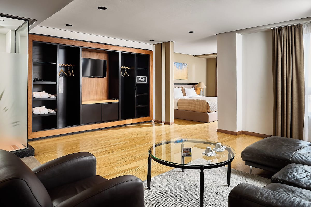 Ein Hotelzimmer mit einem Bett, einer Sitzecke mit zwei Stühlen, einem Glastisch, einem Kleiderschrank mit darin befindlichen Kleidungsstücken und einem Fernseher.