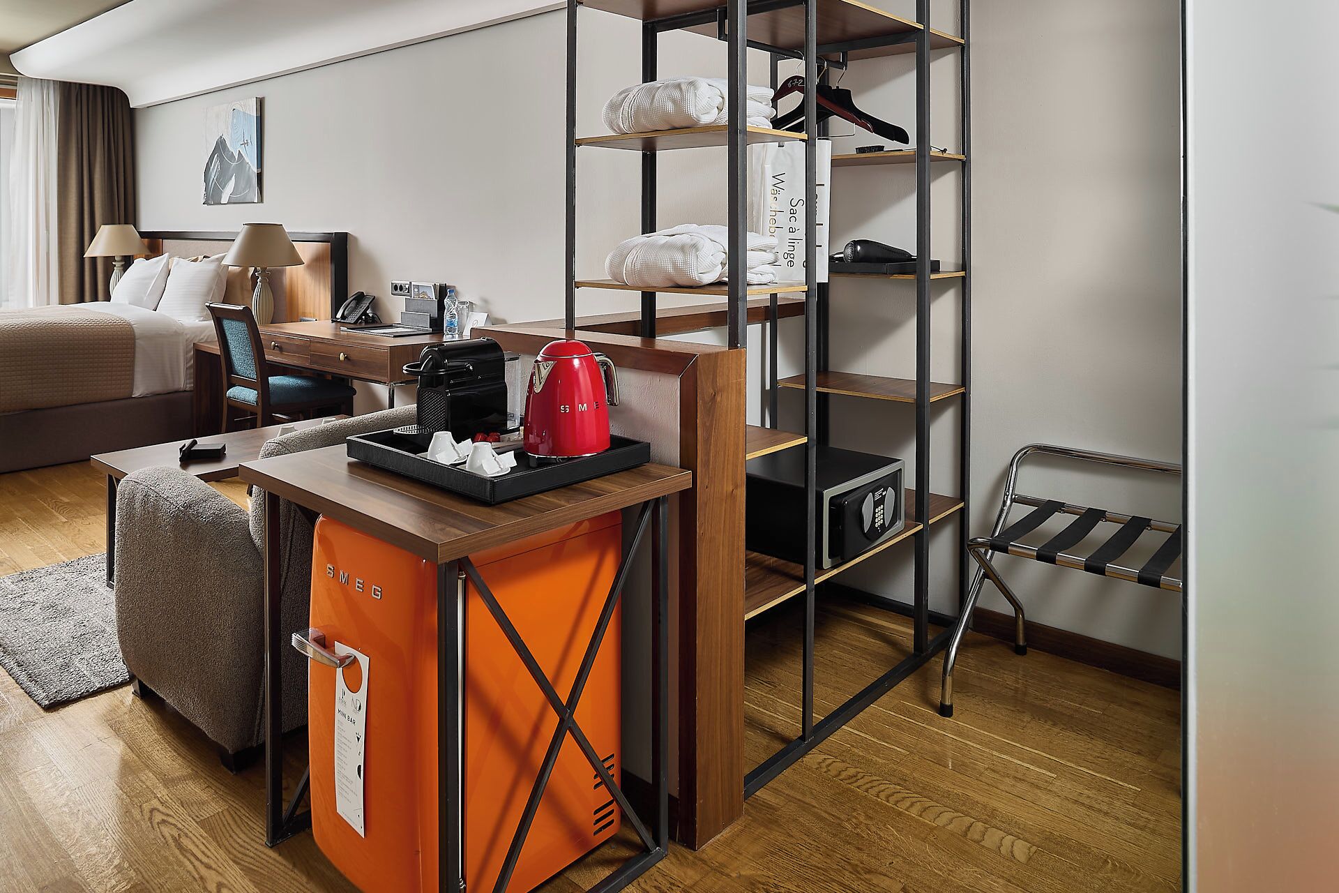 A compact hotel room with a bed, a desk with a coffee maker, a shelf with towels, and a shoe rack.
