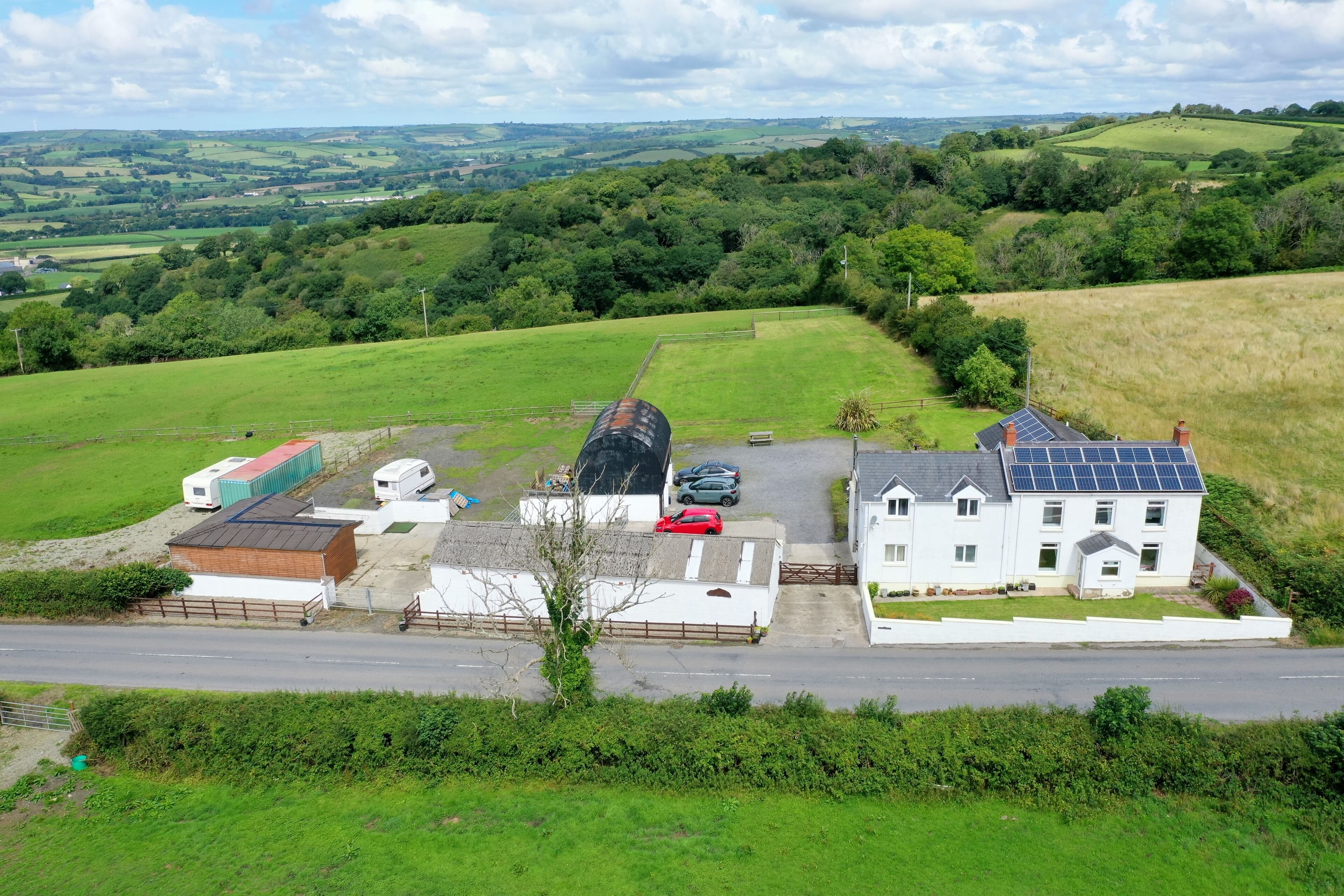 A rural setting with a white house featuring solar panels, a red car, and a trailer park.