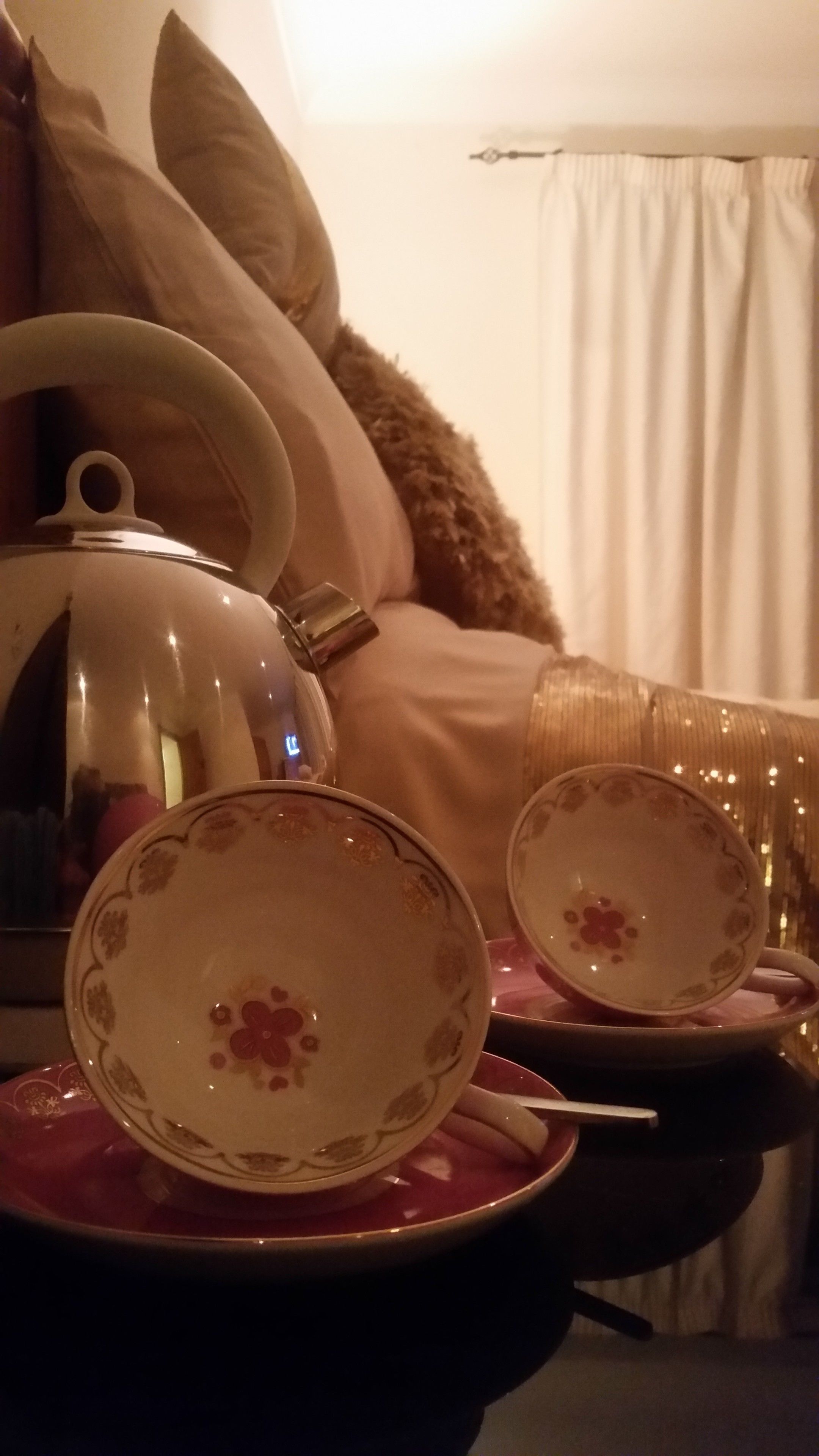 Un jeu de te amb tetera i tasses damunt d'una taula, un llit amb almoades, i una finestra amb cortines.