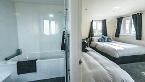 Et hotelværelse med bruseniche i glas, to senge med hvide sengetæpper, og et vindue med gardiner.