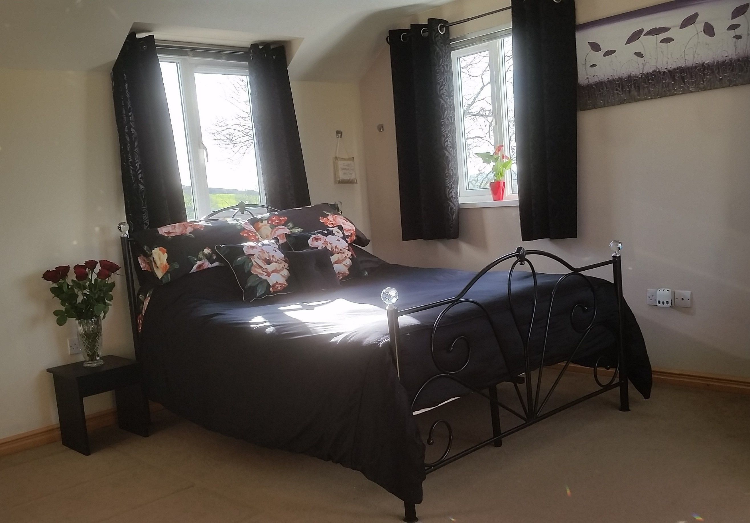 Une chambre à coucher avec un lit en métal noir, une couvre-lit noire et un vase contenant des roses rouges, posé sur une petite table à côté du lit.