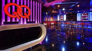 Zona de bar moderna con un llamativo letrero de neón rojo y morado, suelo negro pulido y una barra con taburetes.
