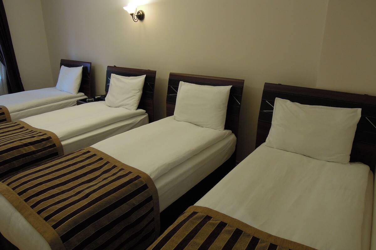 Une chambre d’hôtel avec trois lits simples, chacun doté d’une tête de lit en bois et de draps blancs. On peut voir un luminaire fixé au mur au-dessus des lits.