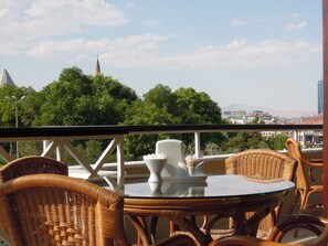 Un balcón con sillas de mimbre y una mesa de vidrio, con vistas a un paisaje urbano que incluye árboles y una torre.