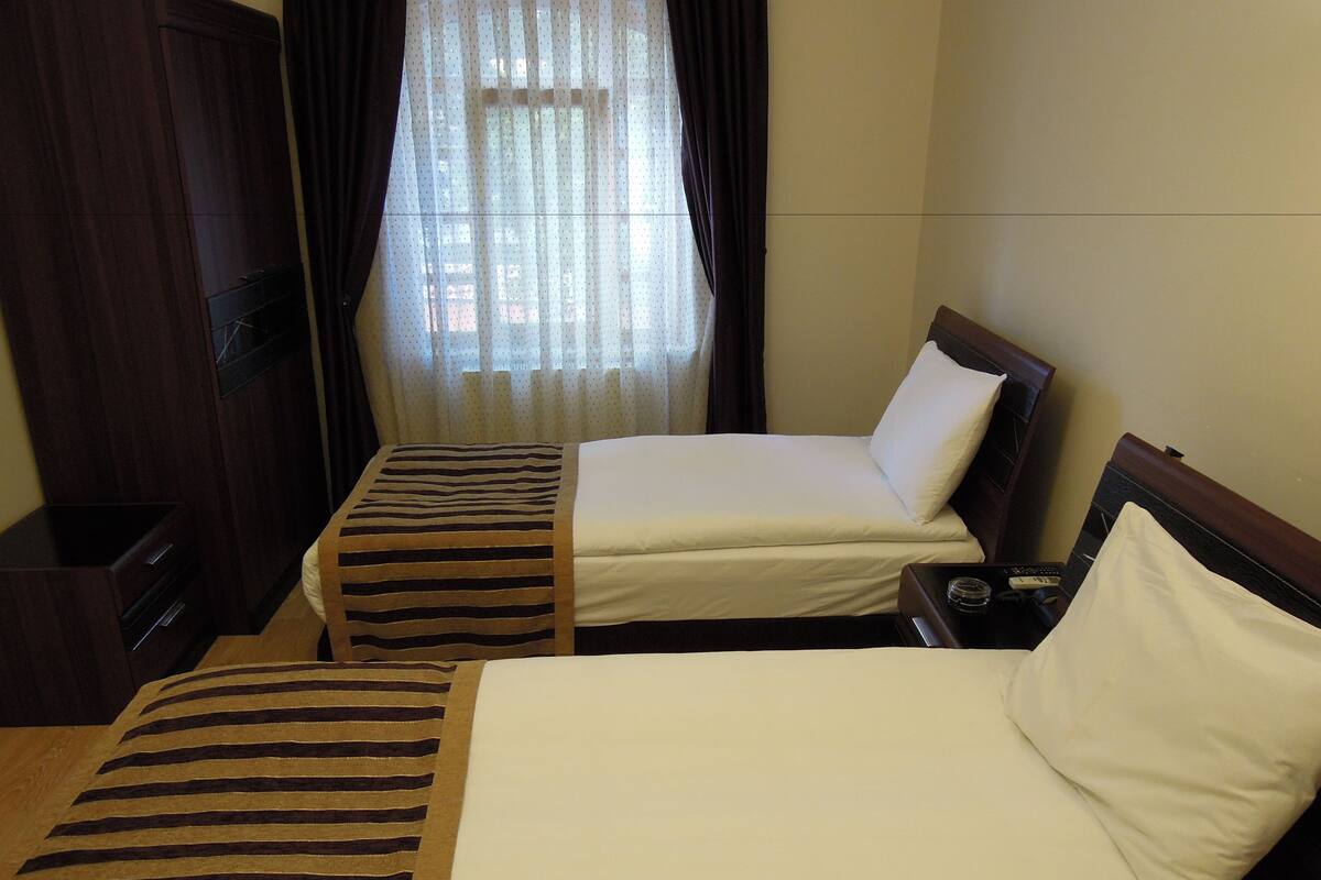 Une chambre d’hôtel avec deux lits, une armoire en bois, une petite table avec une lampe et une fenêtre avec des rideaux légers.