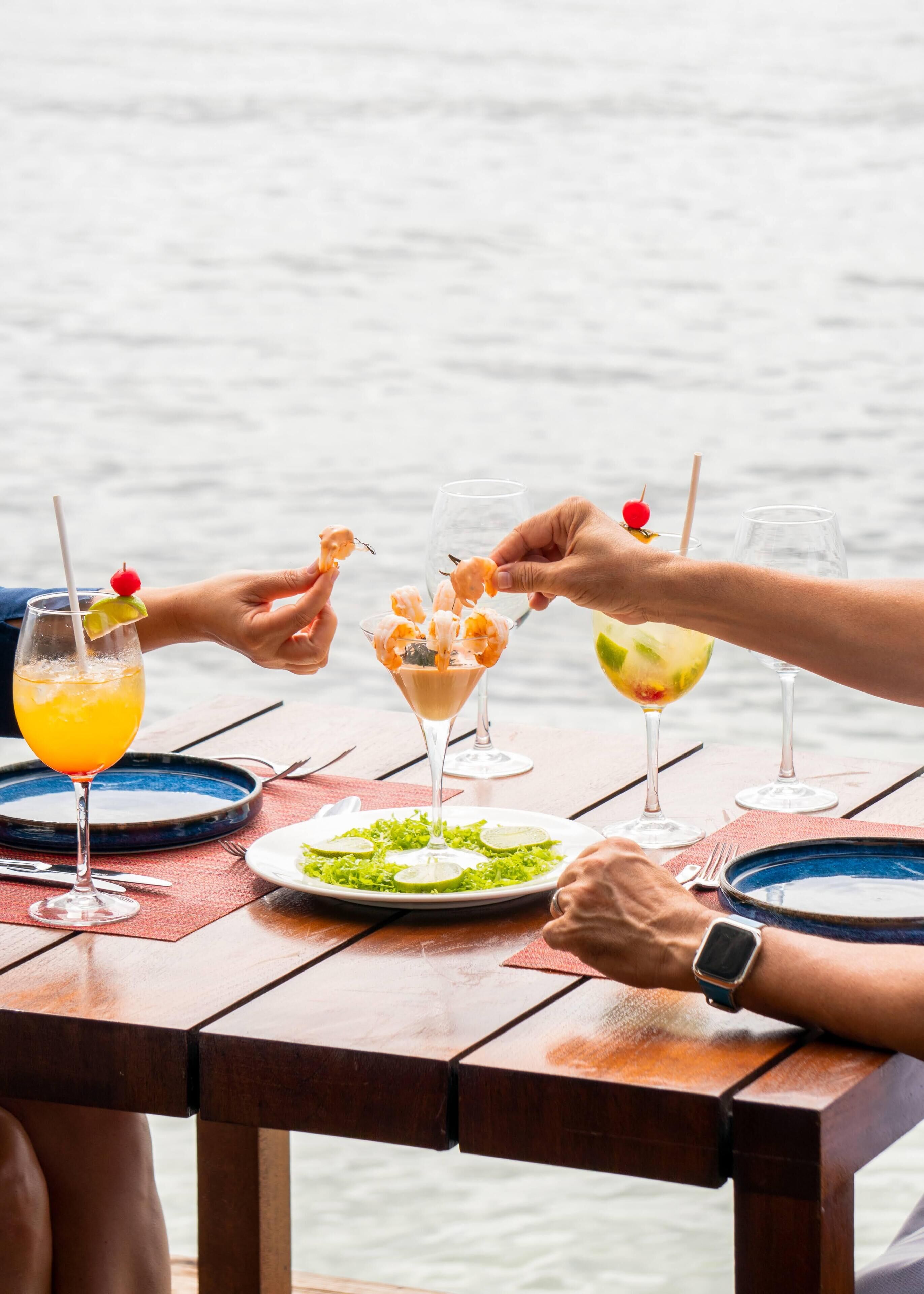 Una mesa de madera con un vaso de jugo de naranja, una copa de martini con camarones y una copa de cóctel con una rodaja de lima.