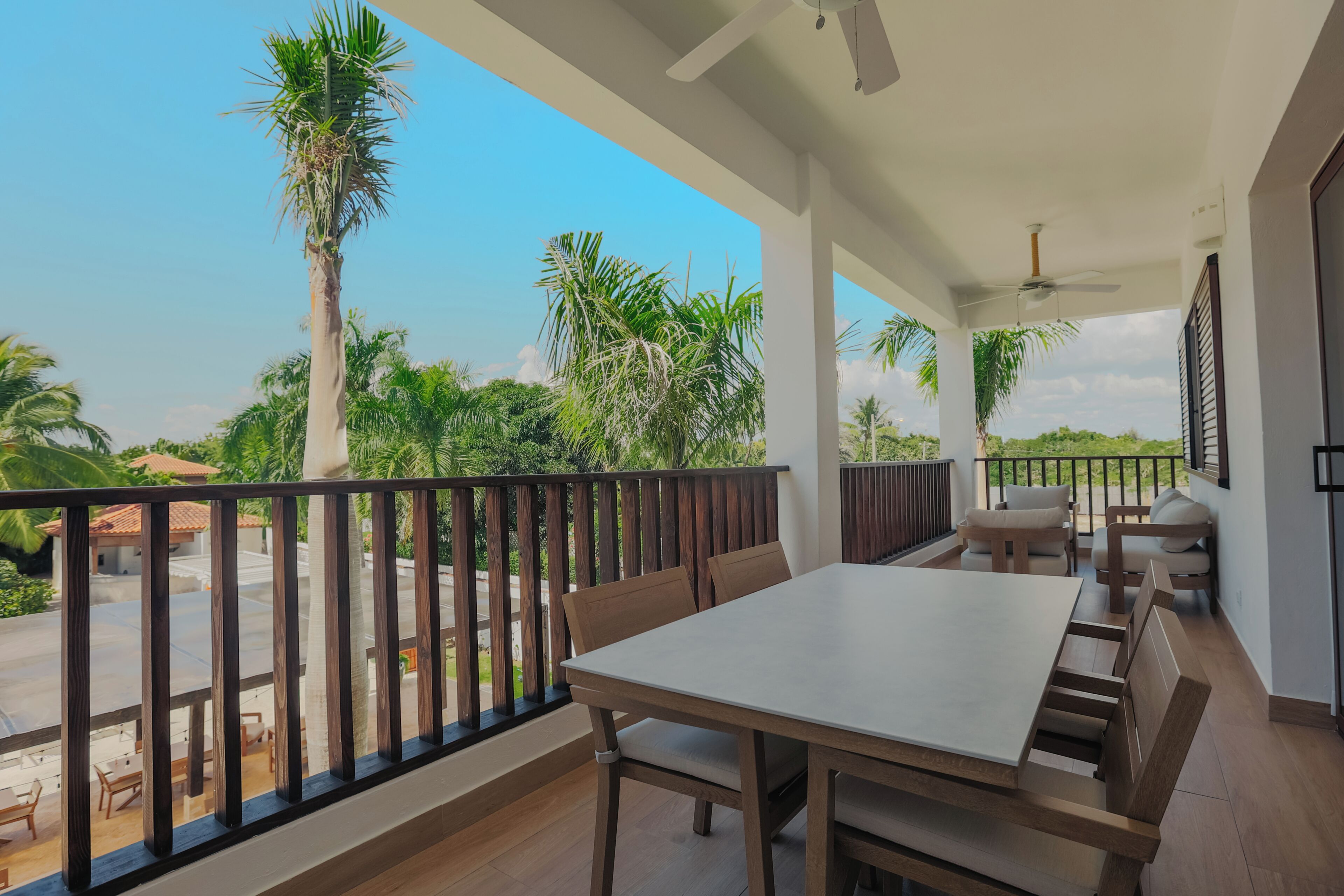 Een balkon met een tafel en stoelen, een plafondventilator en uitzicht op palmbomen en groen.