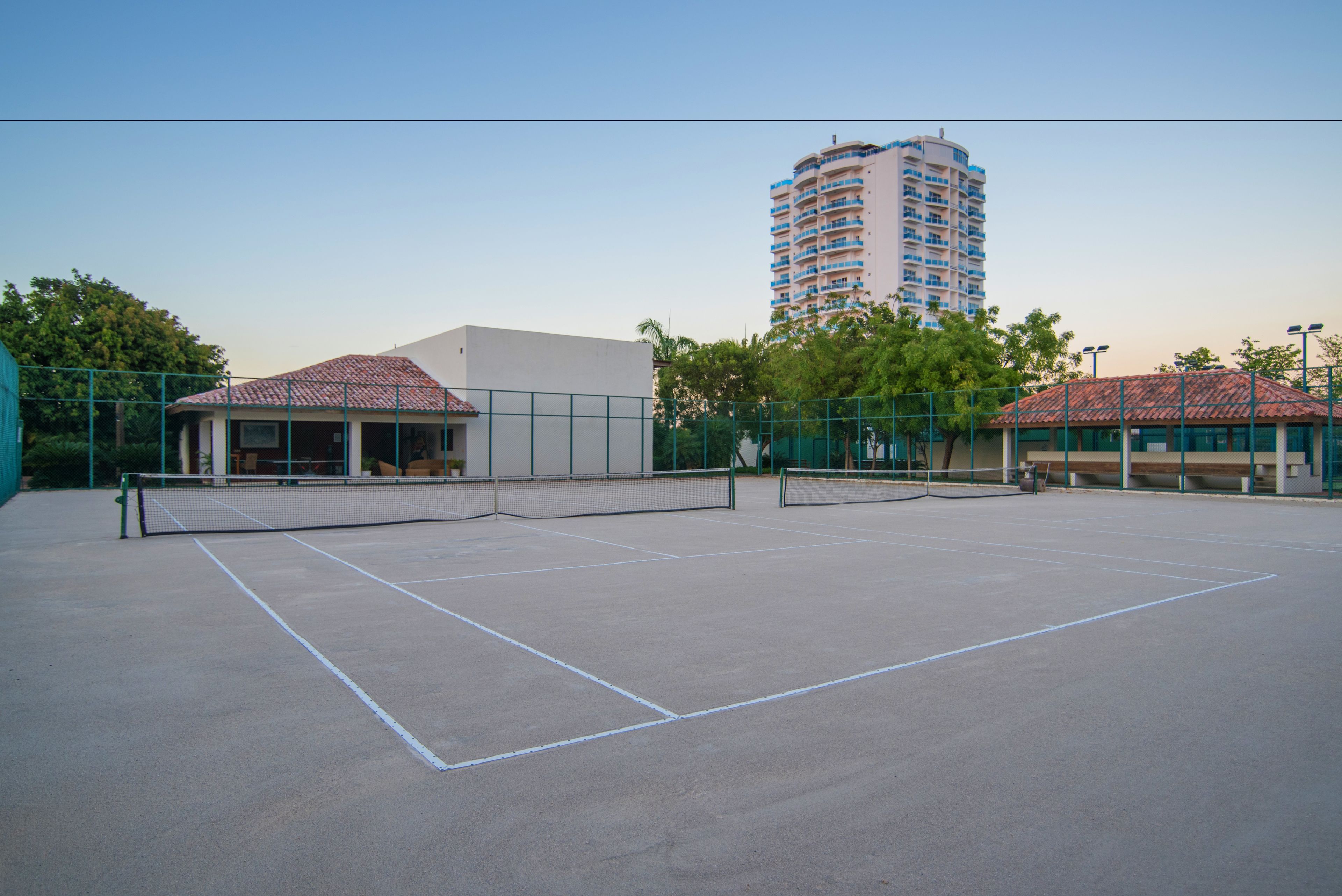 Una cancha de tenis al aire libre con red, rodeada de vallas verdes y edificios con tejados rojos.