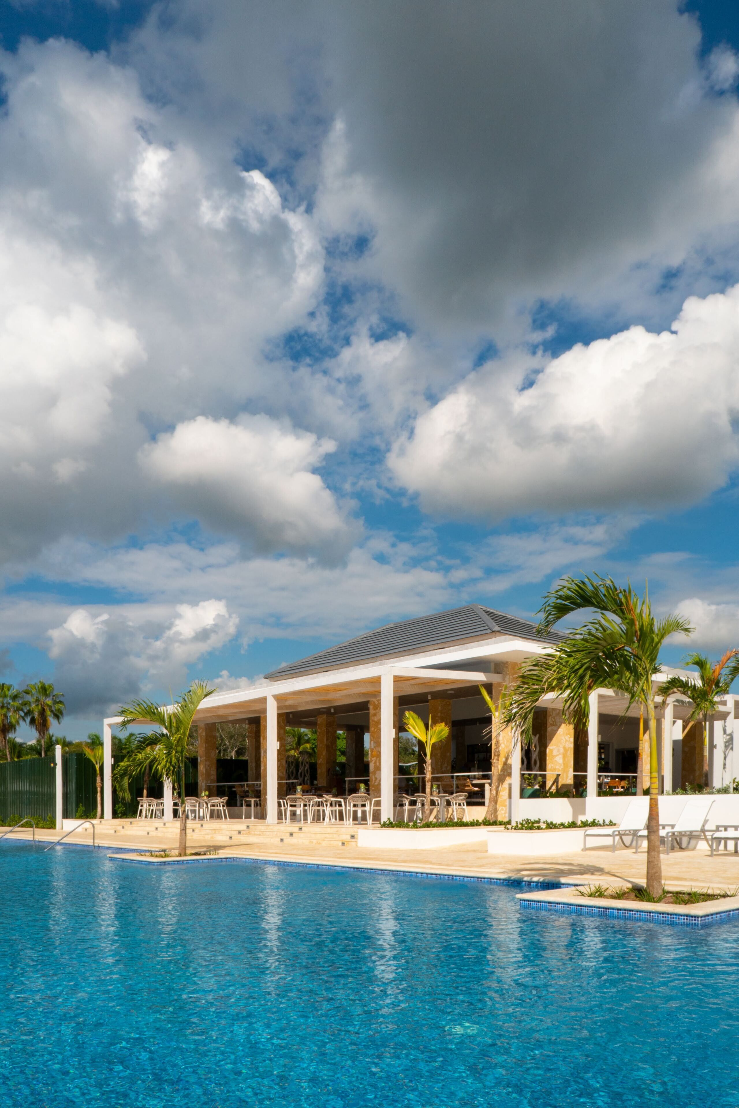 A poolside area with a covered seating area, palm trees, and a clear sky.