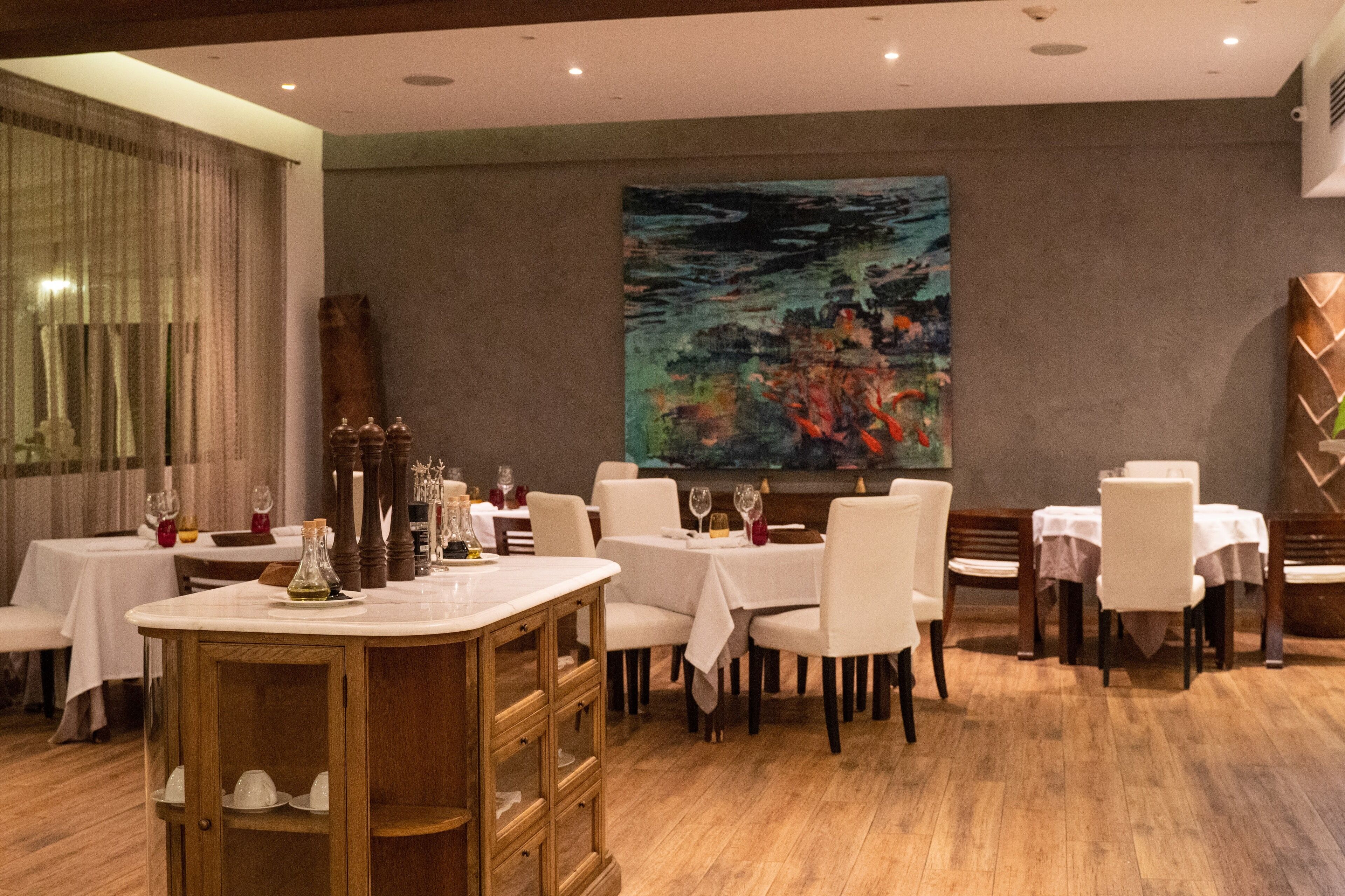 A dining area with round tables, white chairs, and a large abstract painting on the wall.