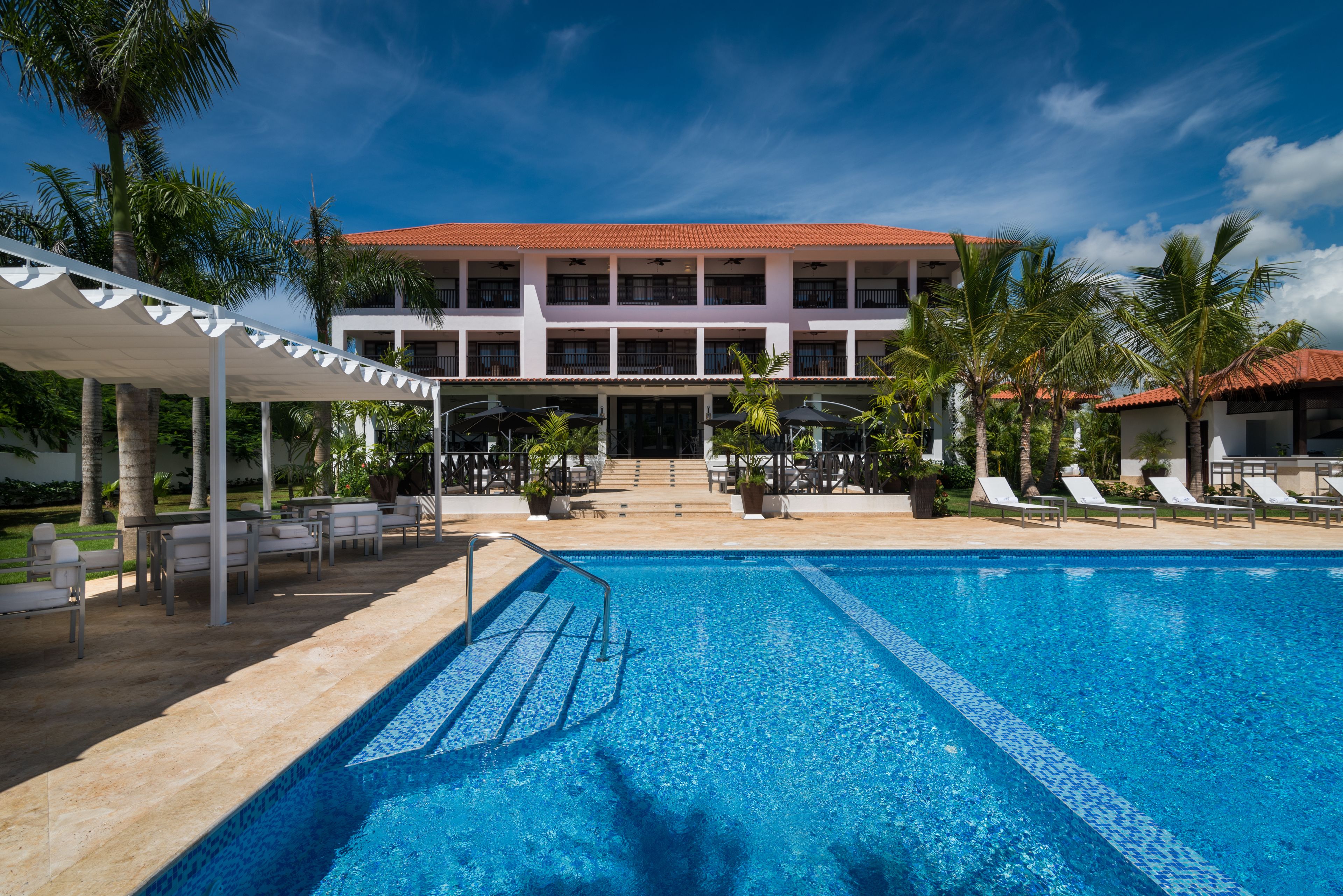 A swimming pool with lounge chairs and umbrellas, a covered seating area, and a building with a red-tiled roof.