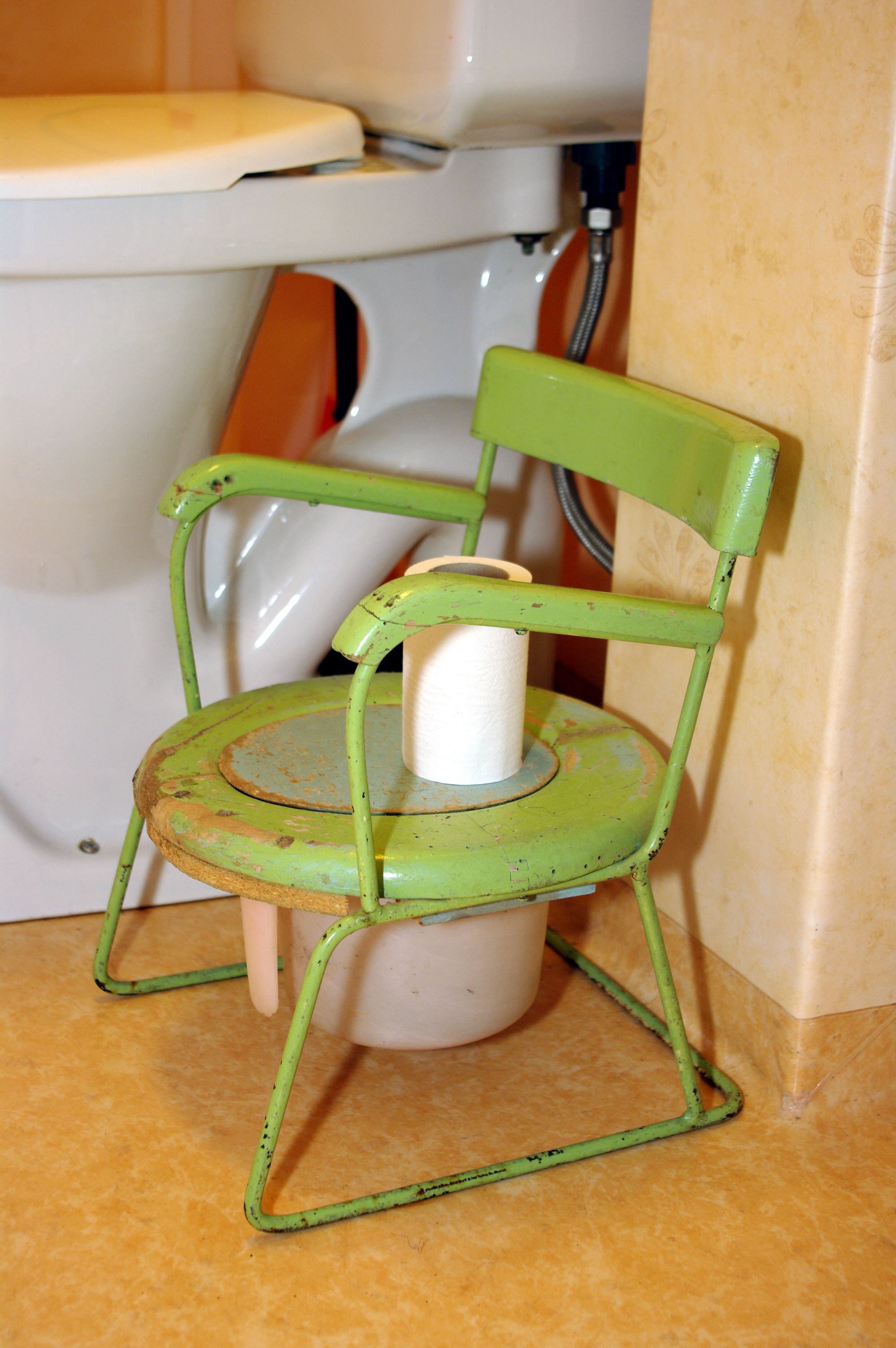 Una silla metálica verde con un asiento de inodoro blanco encima, ubicada en un baño.