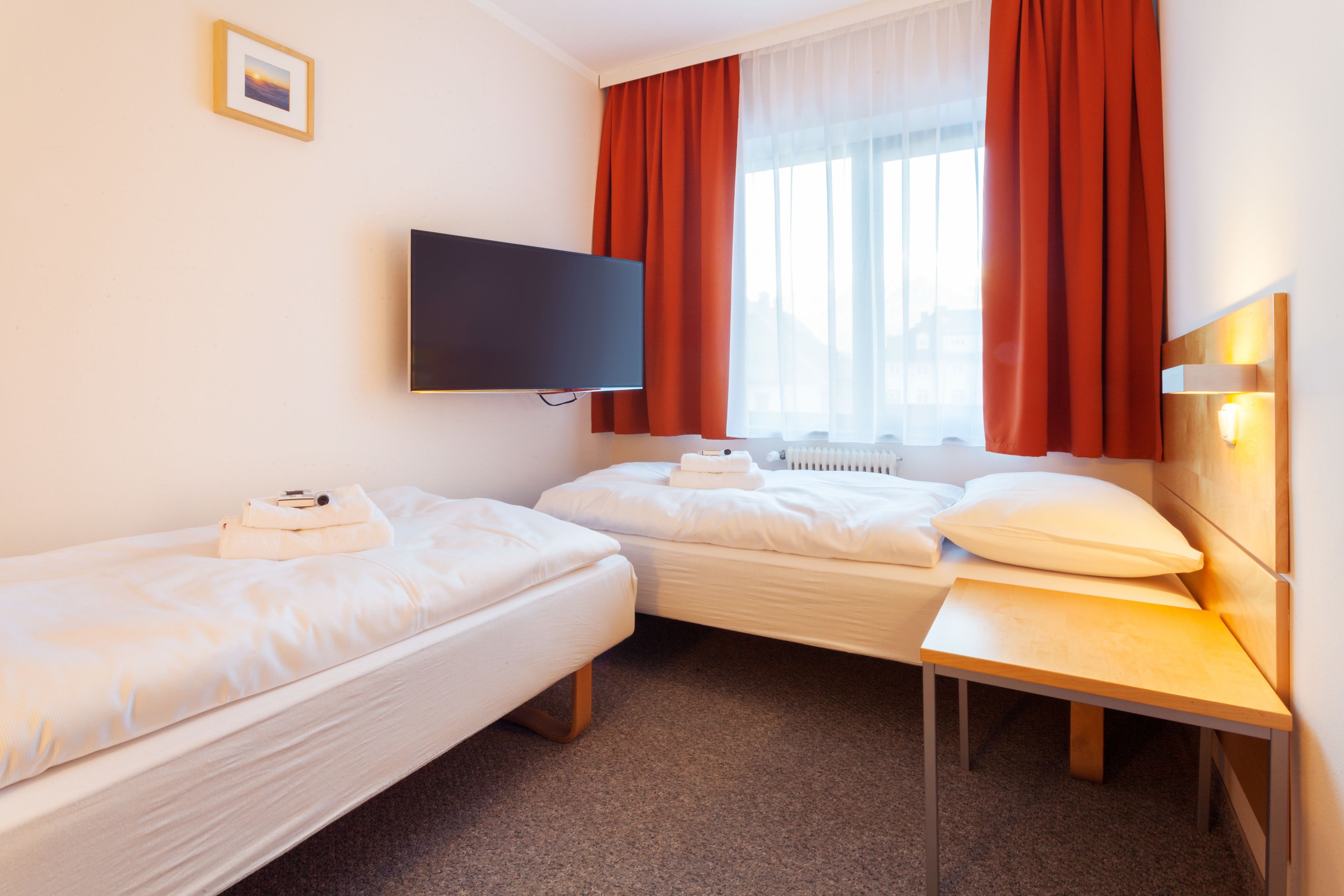 Habitación de hotel con dos camas individuales, un televisor de pantalla plana montado en la pared, una mesita de noche de madera y una ventana con cortinas rojas.