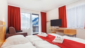 A hotel room with two beds, a desk, a chair, a TV, and a view of mountains through the window. - Residence AlpenHeart (Bad Gastein)