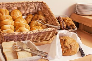 Un panier en osier rempli de différents types de pains et de petits pains, une miche de pain posée sur une planche à découper et une pile d’assiettes blanches.