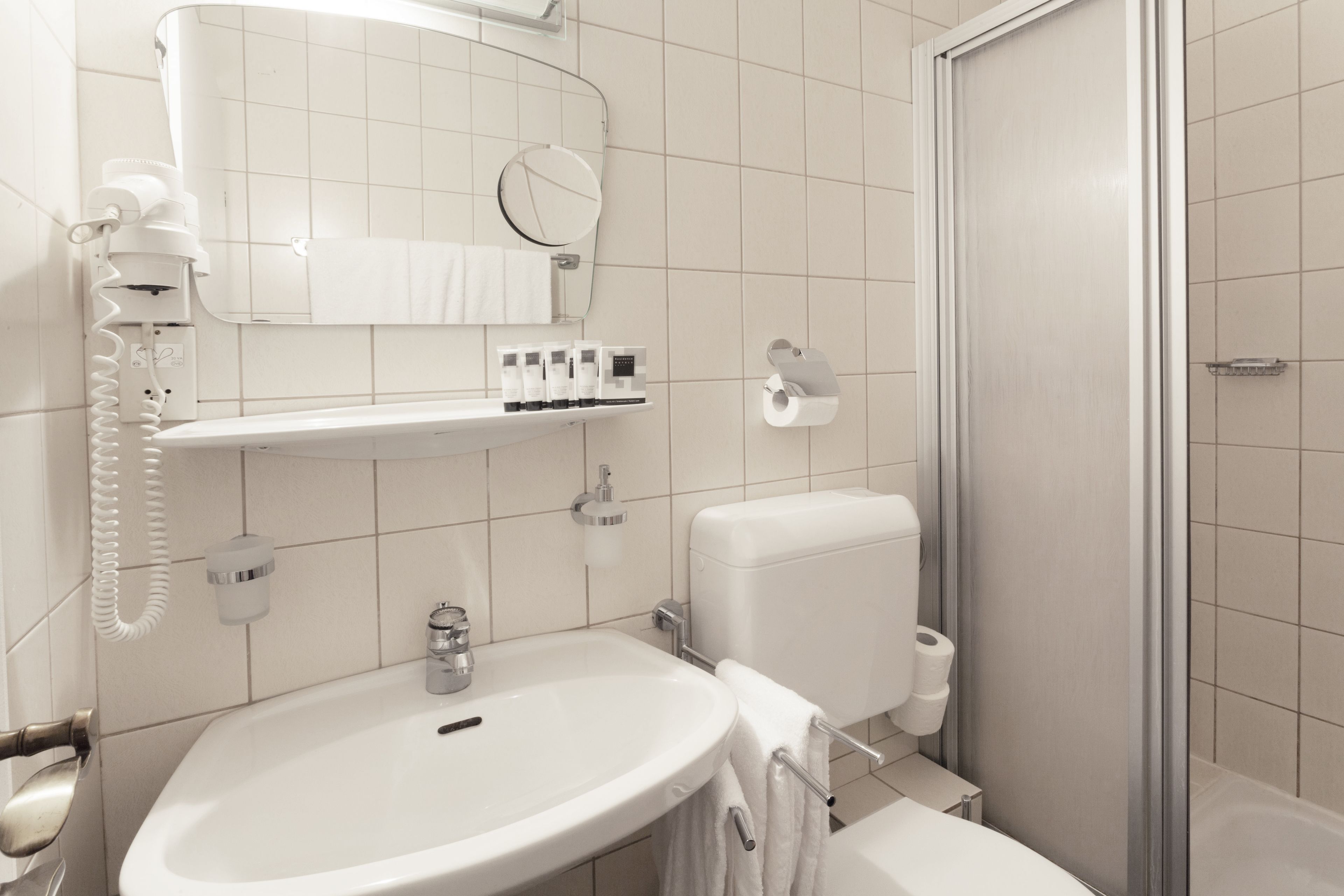 A modern bathroom with a white sink, toilet, and shower. There is a towel rack, a mirror, and a shelf with toiletries.