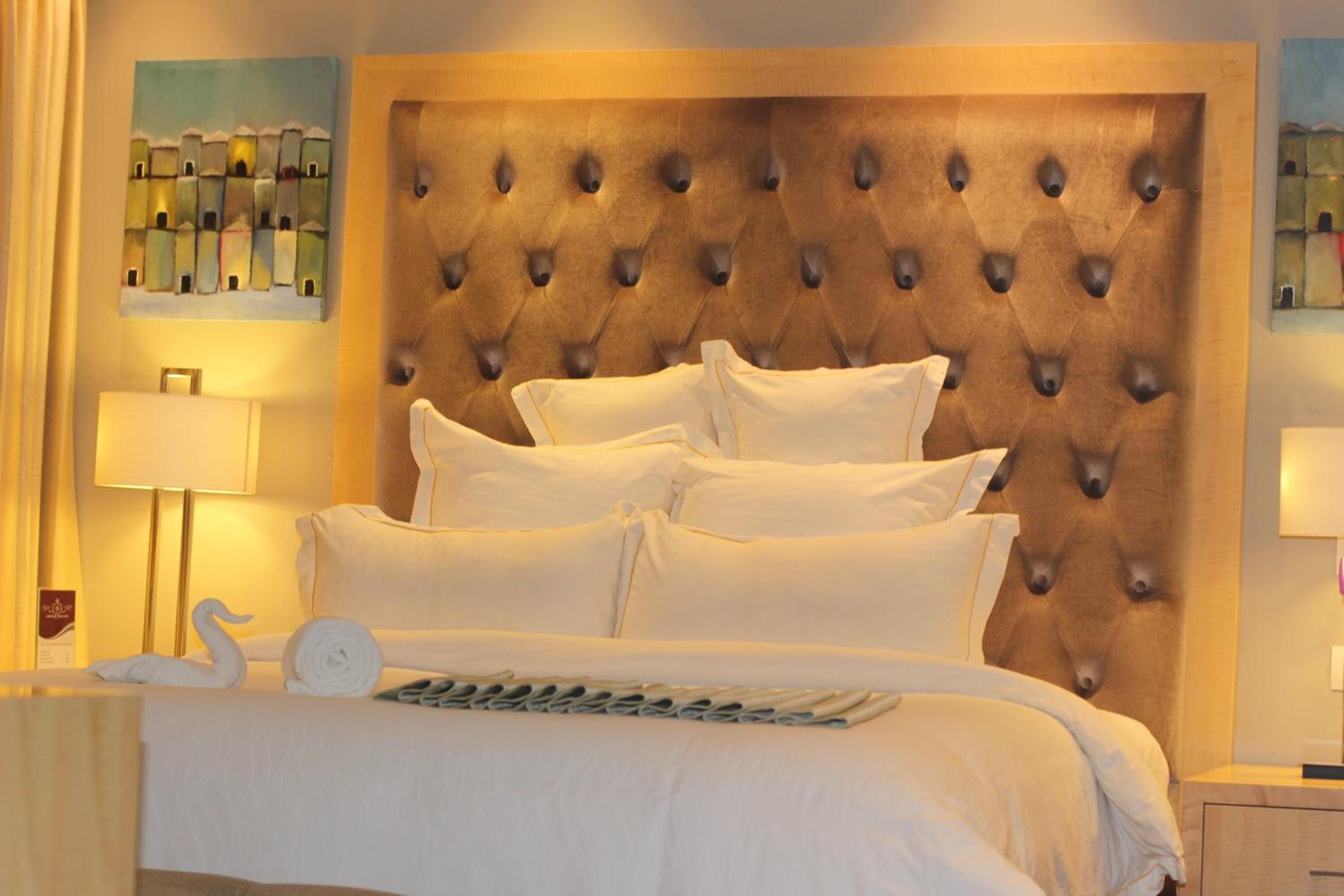 A hotel room with a tufted headboard, a neatly made bed with white linens, a swan-shaped towel, and a wall-mounted lamp.