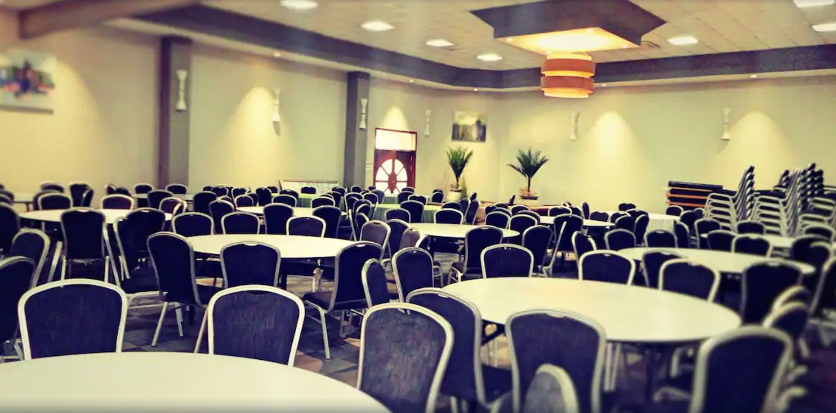 A conference room with multiple round tables and chairs, a stage with a podium, and potted plants.