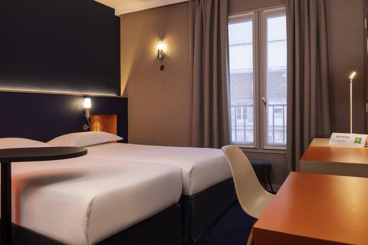 A hotel room with two beds, a desk, a chair, a window with curtains, and a wall-mounted light.