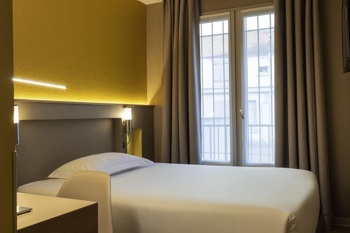Una habitación de hotel con una cama, un escritorio, una lámpara y un ventanal con cortinas.