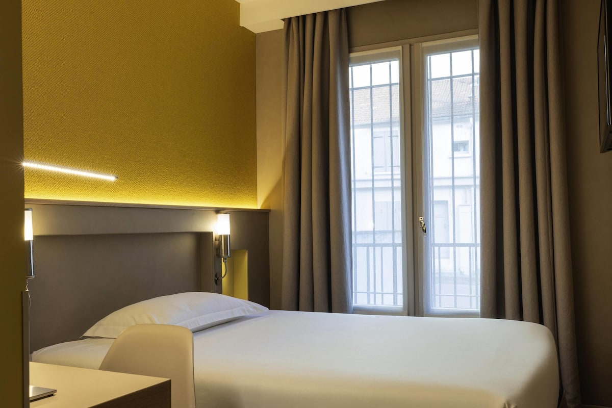 A hotel room with a bed, a desk, a lamp, and a window with curtains.