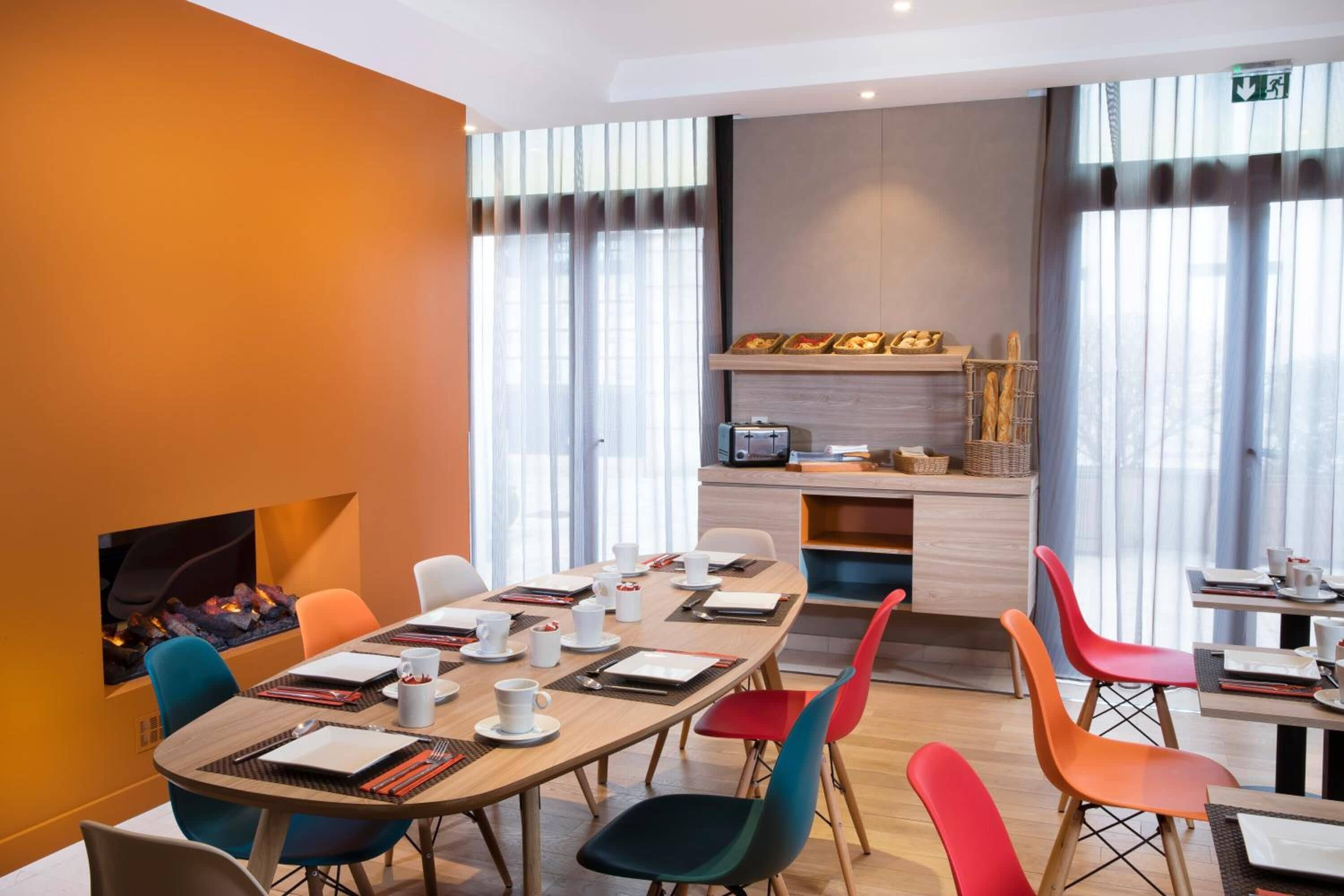 A modern dining area with a long table, colorful chairs, and a kitchen counter with food items.