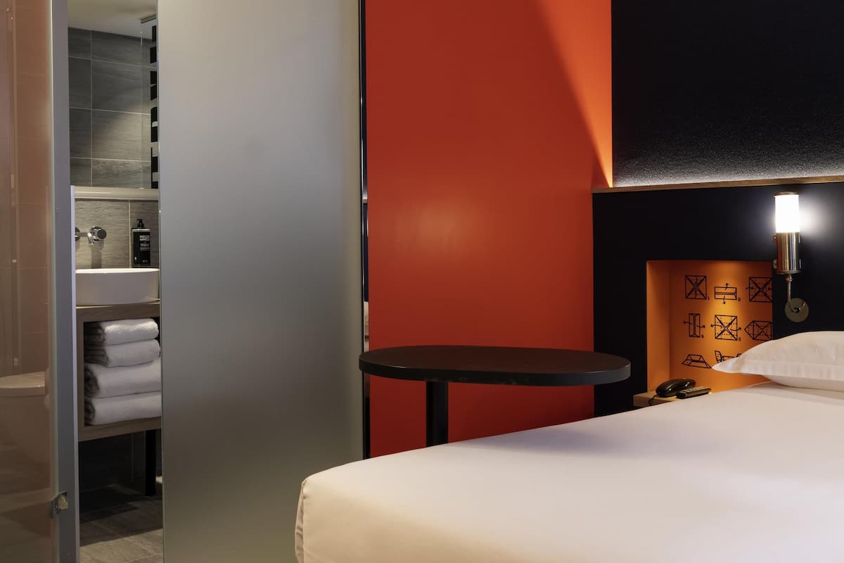 A hotel room with a bed, a small table, a nightstand, a wall-mounted lamp, and a bathroom visible through an open door.