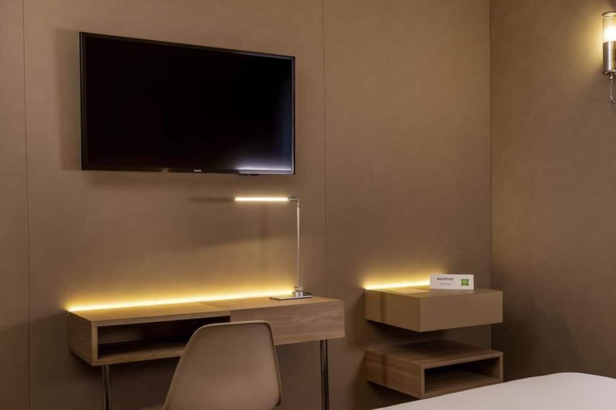 Una habitación de hotel moderna con televisión de pantalla plana montada en la pared, un escritorio con lámpara, una silla y una cama.