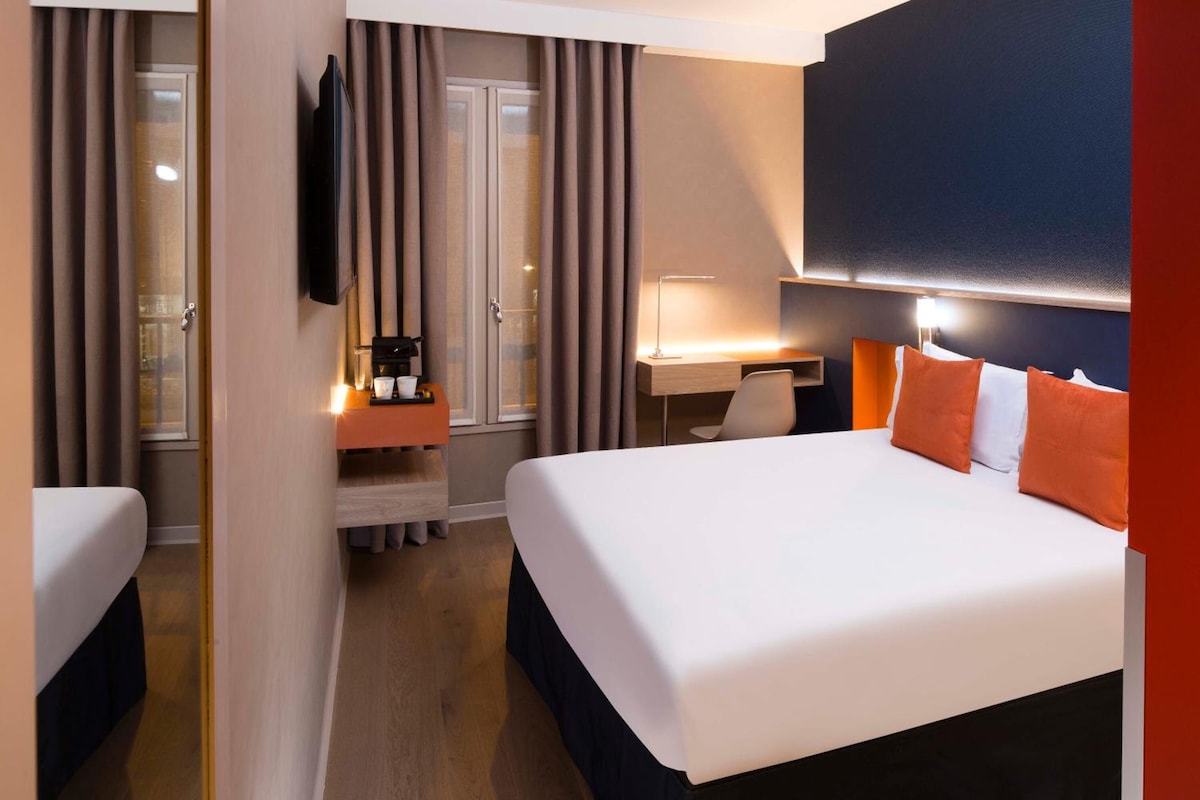 A hotel room with a large bed, a desk, a chair, a TV, and a window with curtains.