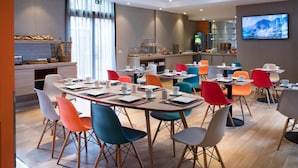 Ein modernes Hotelrestaurant mit mehreren Tischen und bunten Stühlen, einer Küche im Hintergrund und einem an der Wand befestigten Fernseher.