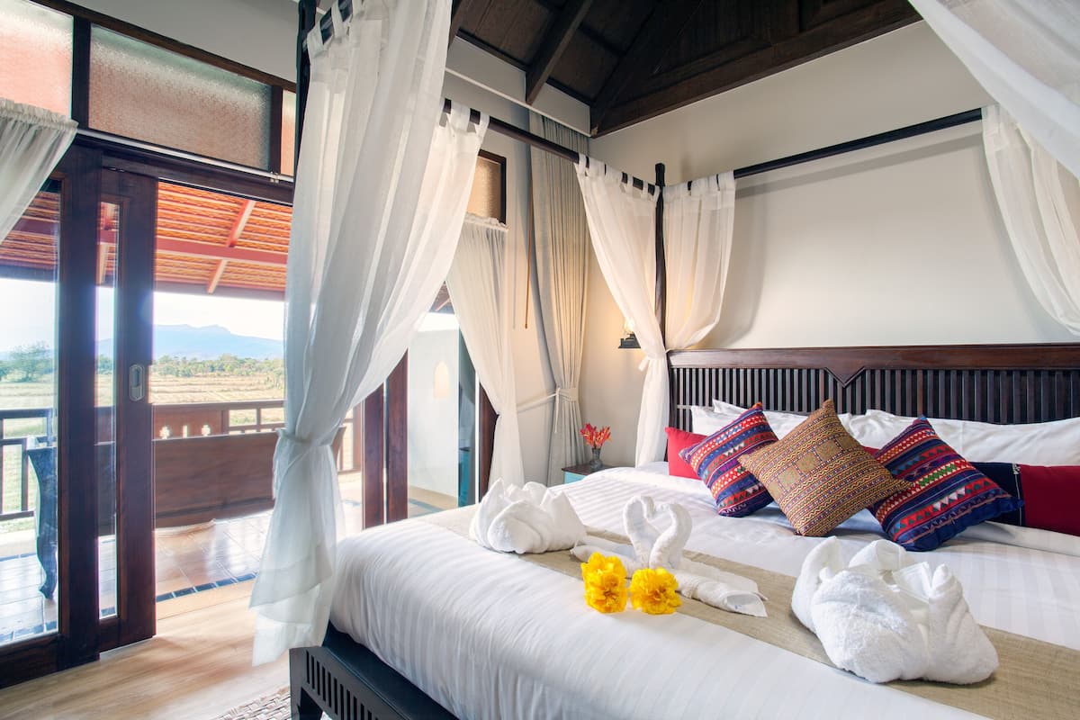A hotel room with a bed, white curtains, a wooden headboard, and a view of the outdoors.