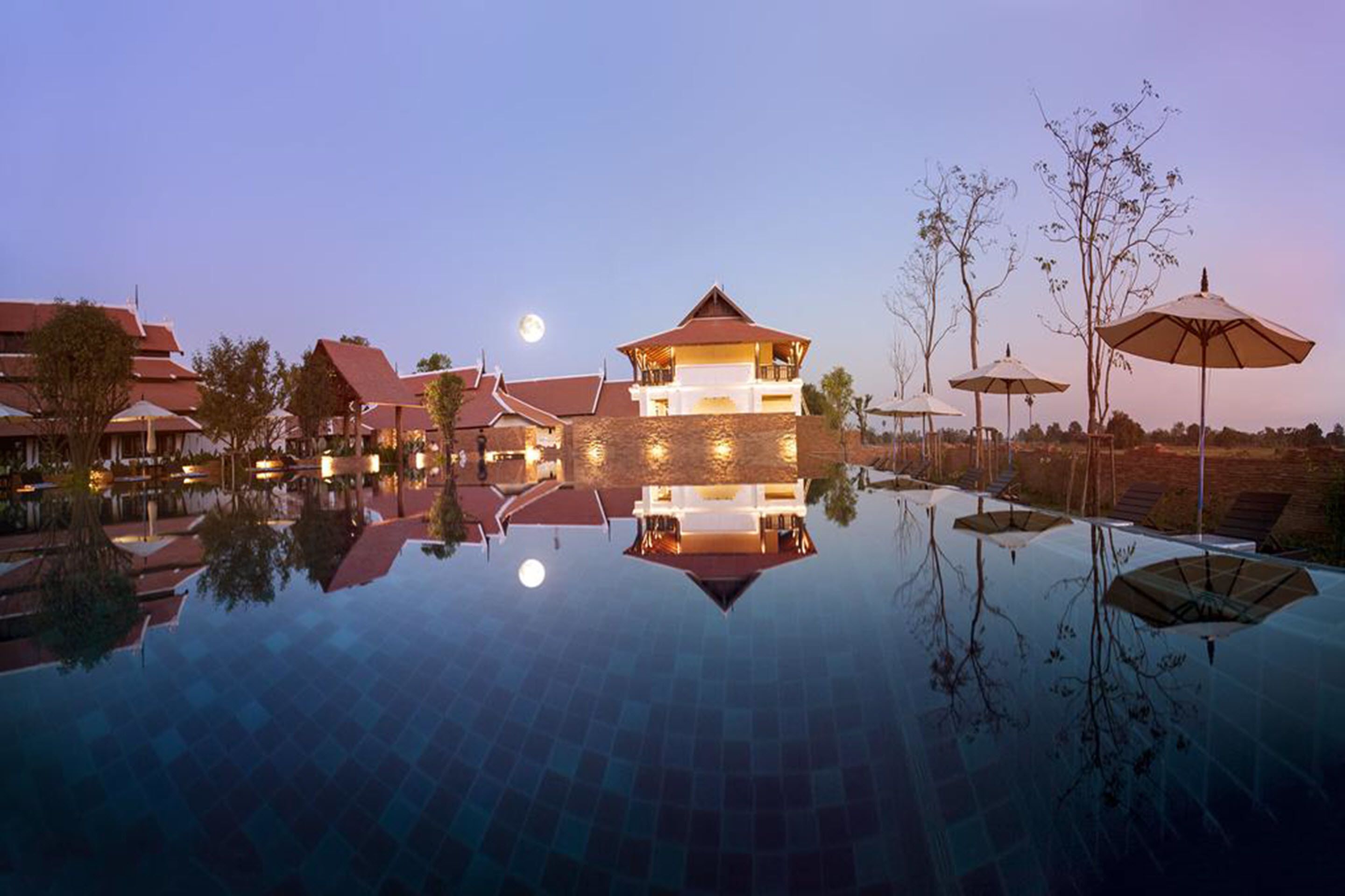 Un hotel con piscina a specchio, un edificio importante con tetto rosso e diversi ombrelloni.