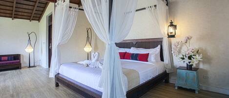 A four-poster bed with white bedding and red pillows, a small table with a vase of flowers, and a bench with cushions in a room with wooden flooring and a high ceiling.