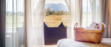 A bedroom with a bed, a wicker basket with towels, and a view of mountains through sheer curtains.