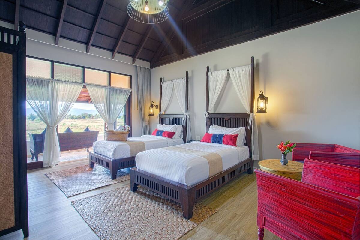 A bedroom with two beds, a red bench, a small table with a vase, and a view of the outdoors.