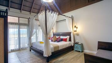 A four-poster canopy bed with a wooden frame and white bedding, a small bedside table with a lamp, and a view of the outdoors through large windows.