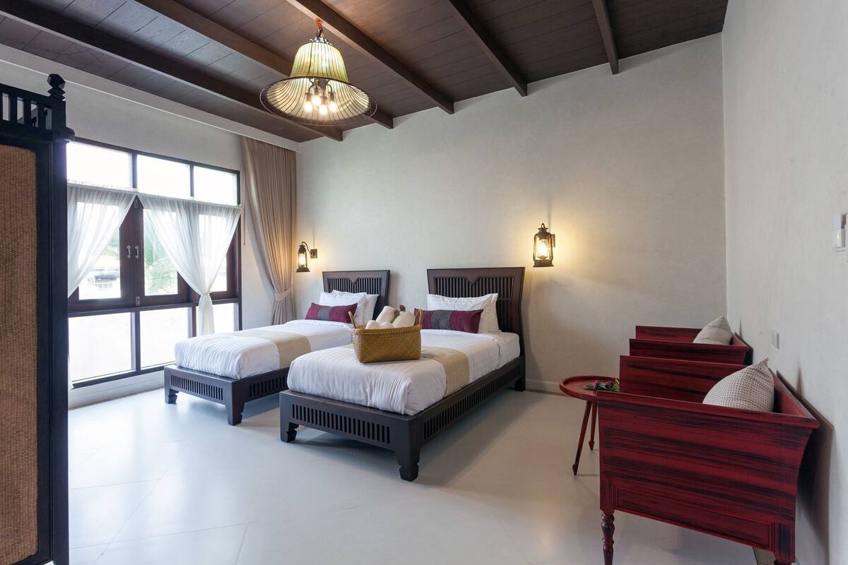 A hotel room with two beds, a wooden bench, a chandelier, and a view of the outdoors.