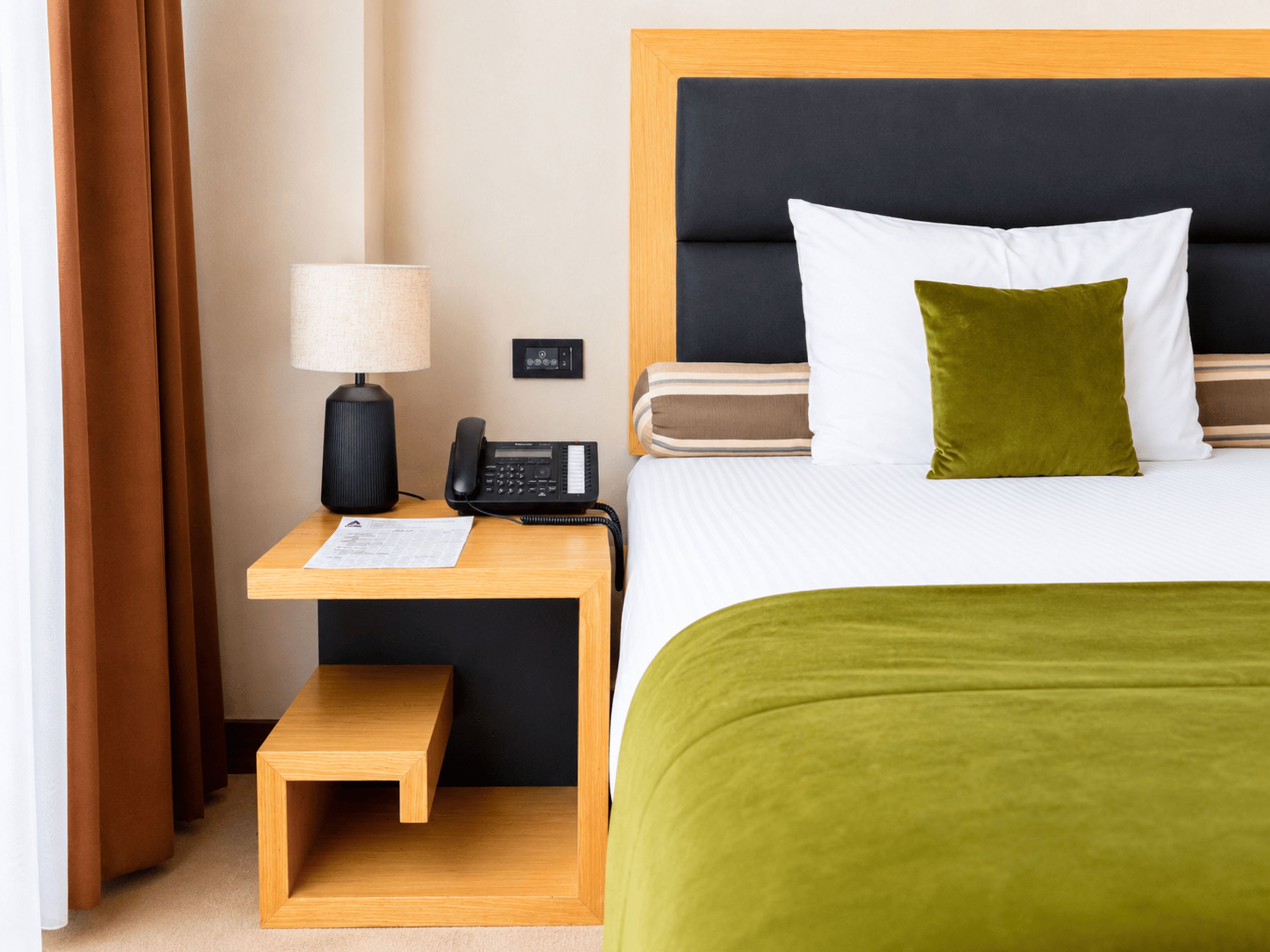 A hotel room with a bed, a bedside table with a lamp and phone, a small wooden stool, and a green throw blanket.