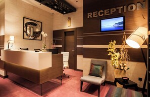 A reception area with a wooden reception desk, a television screen, a mirror, and a chair with a cushion. - Atera Business Suites (Belgrade)