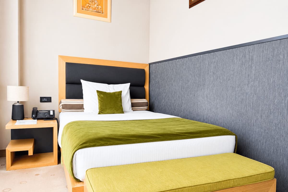 A hotel room with a bed, bedside table, lamp, and a small stool.