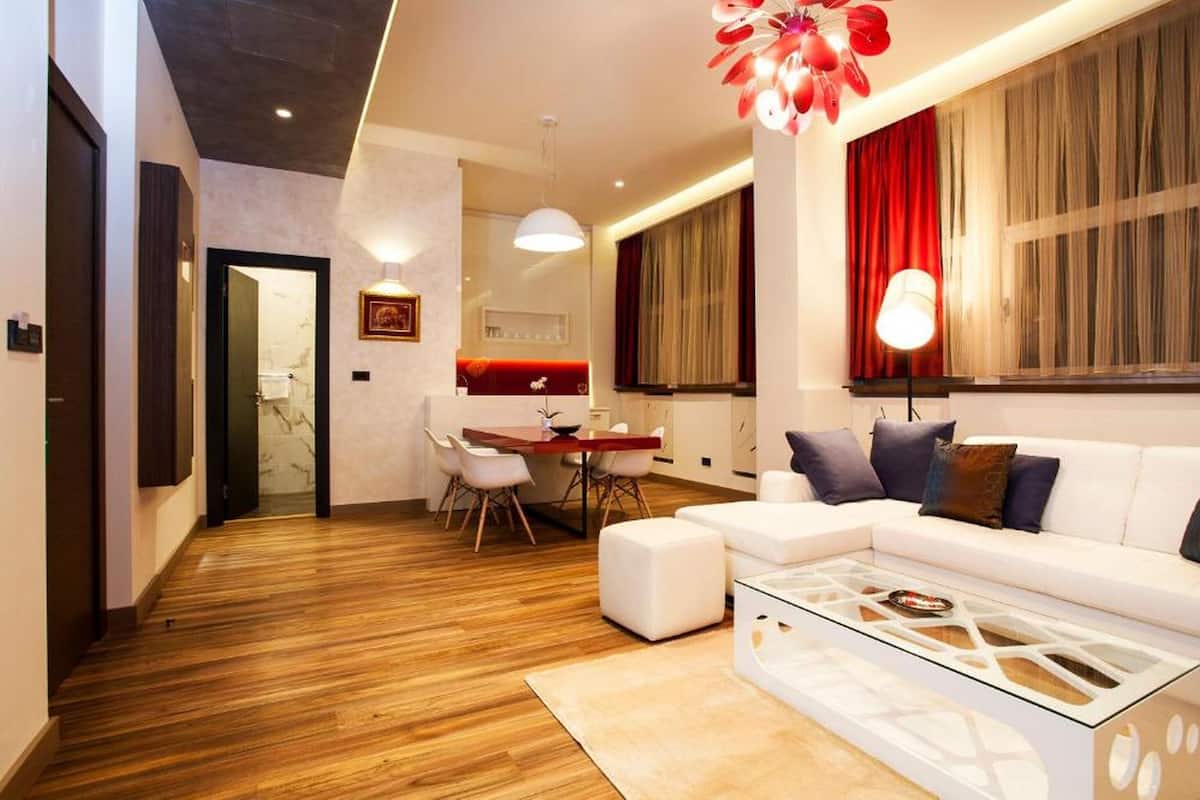A modern living room with a wooden floor, a white sofa, a glass coffee table, and a dining area with a red accent wall.