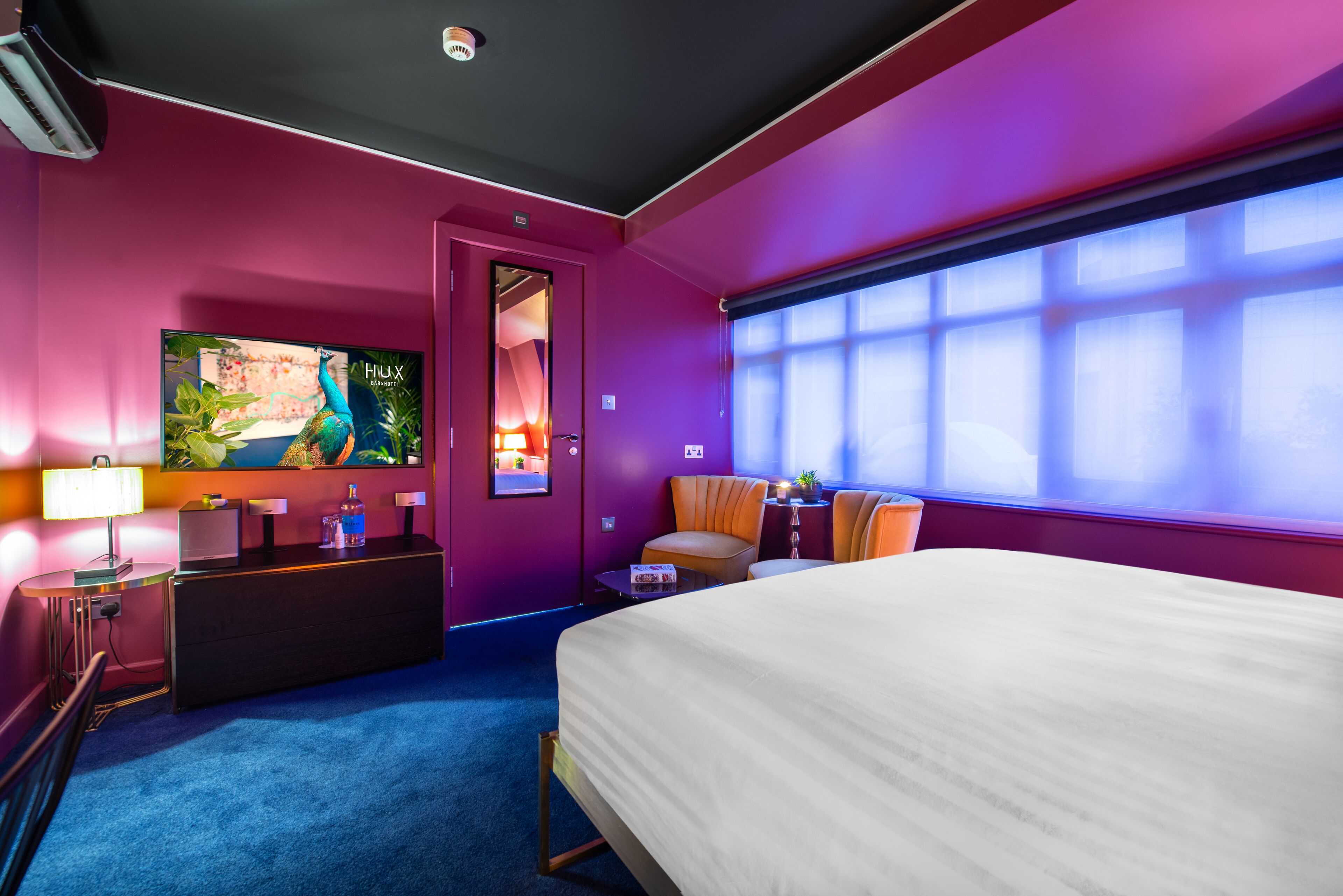 Une chambre d’hôtel avec un grand lit, un bureau avec une lampe, une chaise et une grande fenêtre avec des stores.