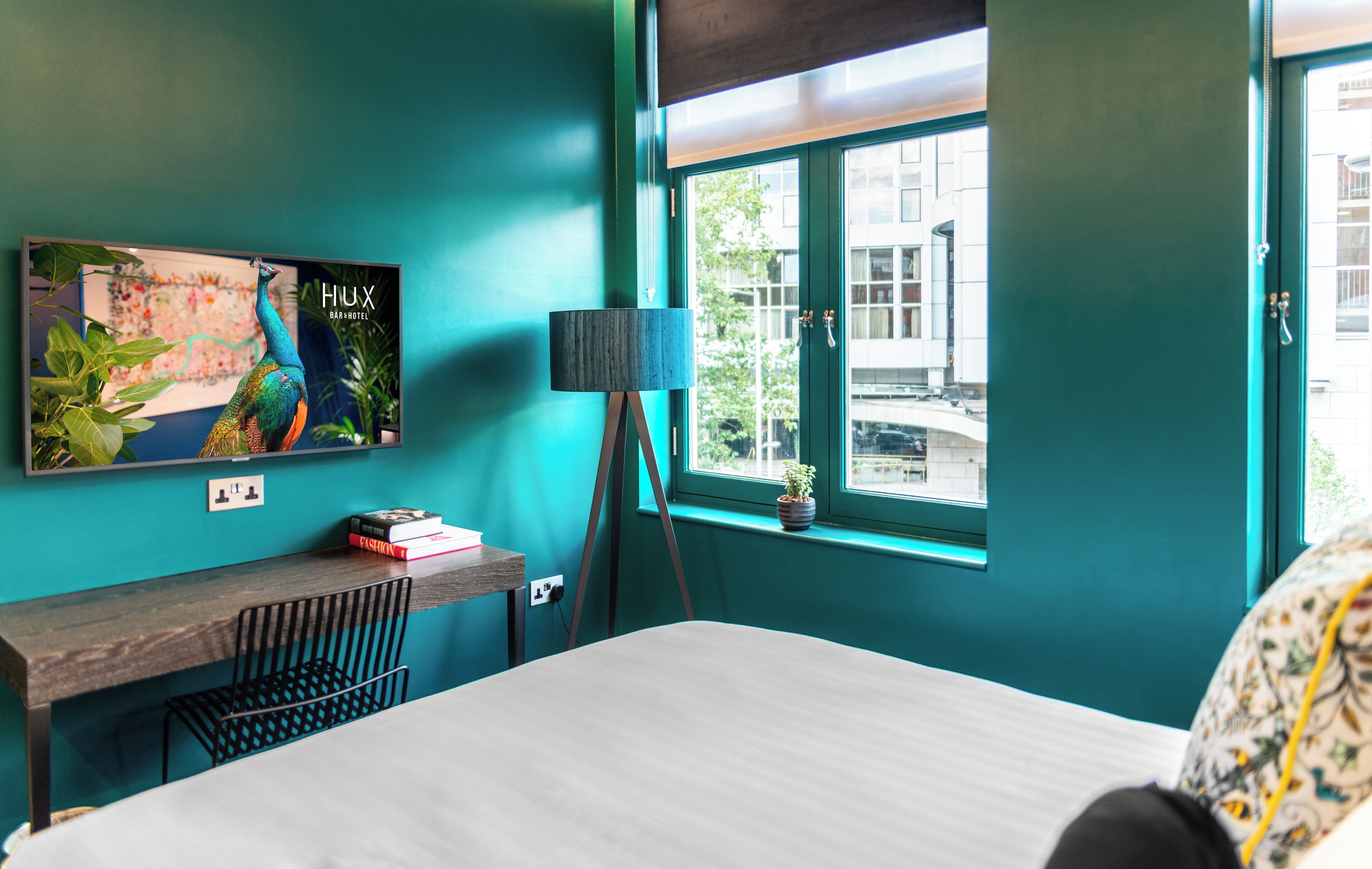 A teal-themed hotel room with a bed, a desk, a lamp, and a window with a view of buildings.