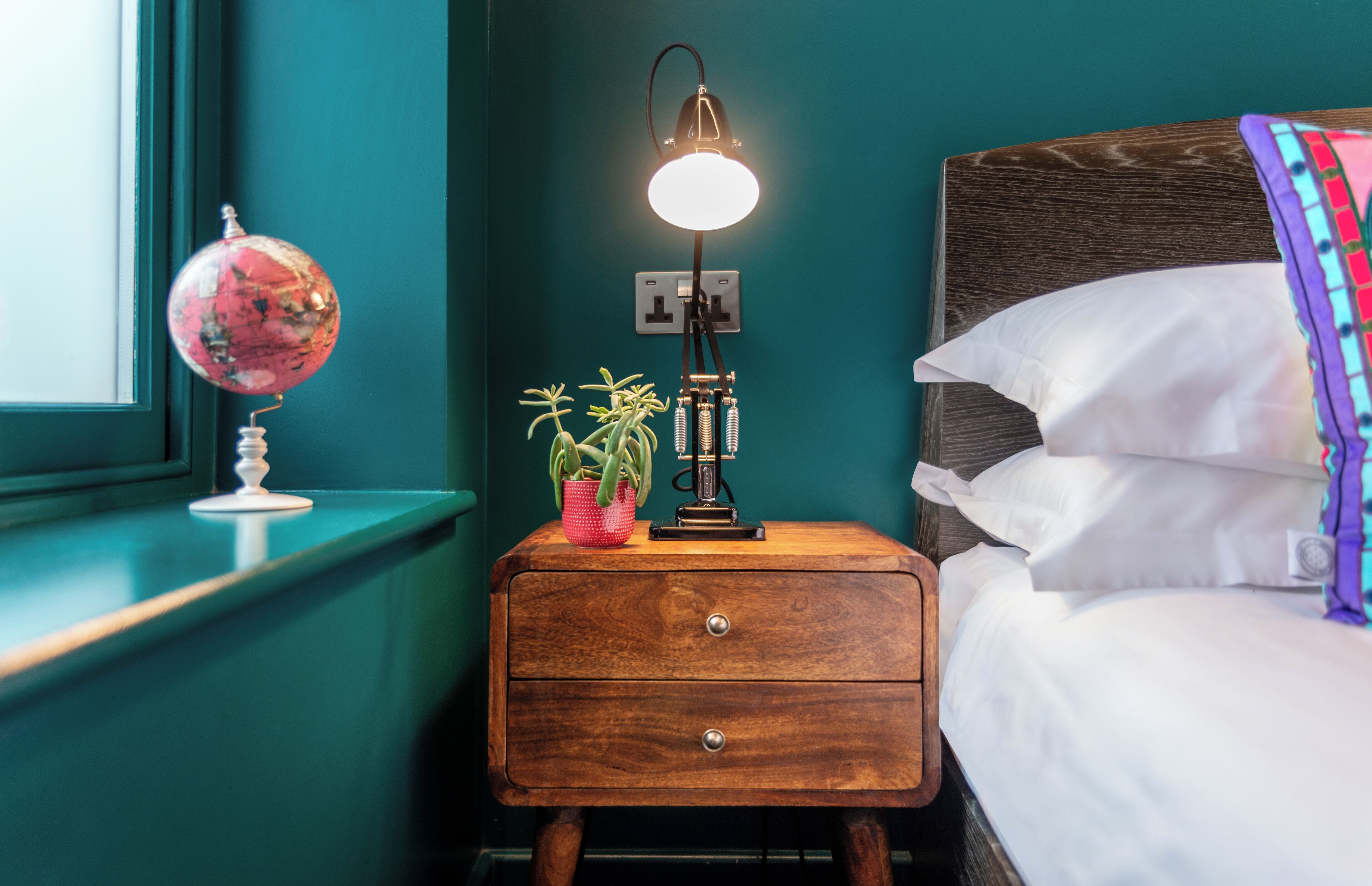 A bedroom with teal walls, a wooden nightstand with a globe and a plant, a bedside lamp, and a bed with white linens and a colorful blanket.