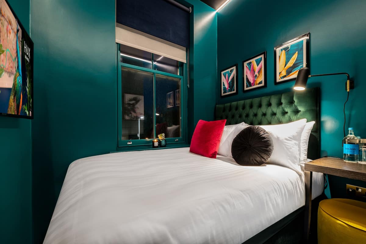 A bedroom with a green upholstered headboard, white bedding, a red pillow, and a bedside table with a lamp and a bottle of water.