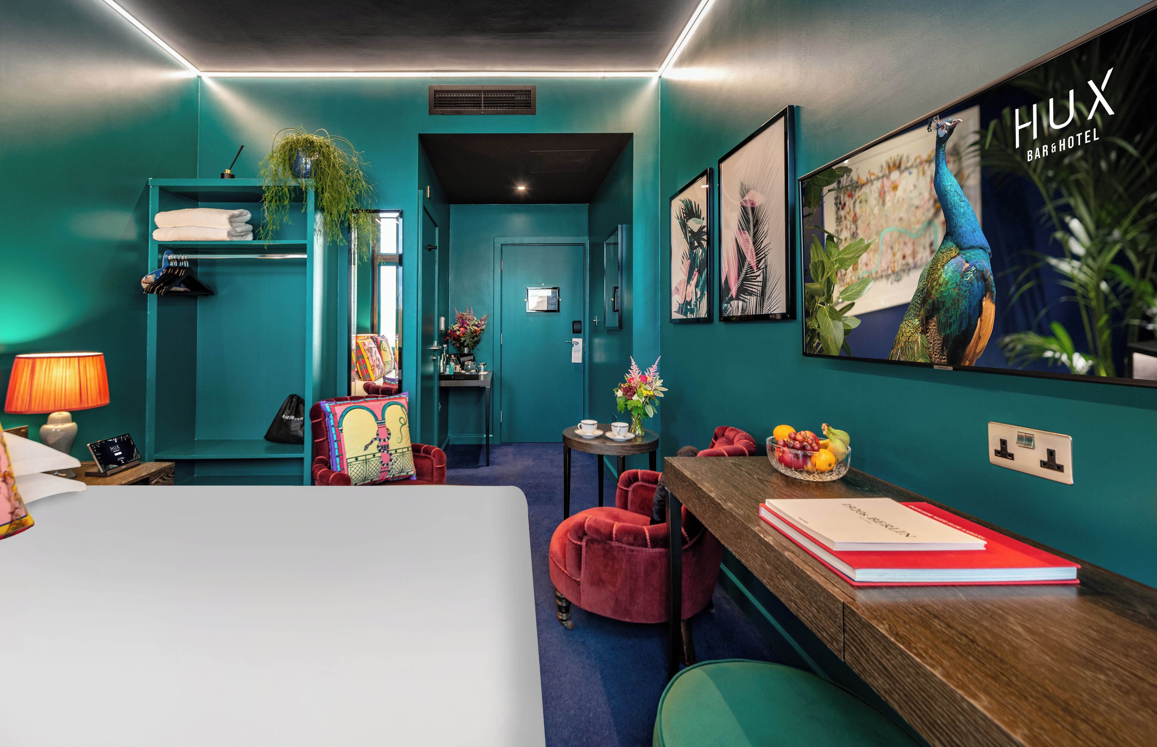 A teal-themed hotel room with a bed, a desk with a red notebook, a red chair, a small table with a vase of flowers, and a wall-mounted TV displaying 'HUX BAR&HOTEL'.