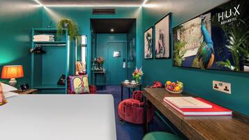A teal-themed hotel room with a bed, a desk with a red notebook, a red chair, a small table with a vase of flowers, and a wall-mounted TV displaying 'HUX BAR&HOTEL'.